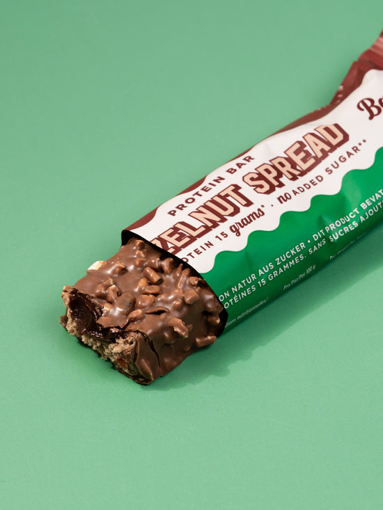 Single Barebells Hazelnut Spread bar with wrapper partially open, revealing chocolate coating with hazelnut pieces on mint background
